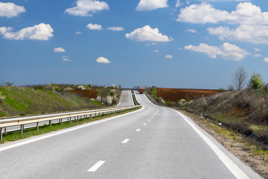 Bulgarian E79 international road as a two-lane single carriageway running through agricultural land between Vidin and Montana, with HGVs cresting a hill in the distance, blue spring sky with scattered clouds, April 2026