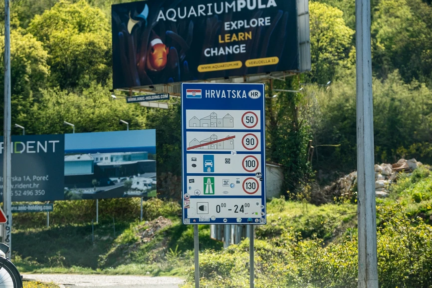 A two-lane road curving towards an international border checkpoint canopy with a distinct blue roof trim on the Slovenia-Croatia border, featuring a motorcycle with two riders traveling in the foreground, with the Slovenian national flag and European Union flag flying on the right side under a clear blue sky, April 2026 