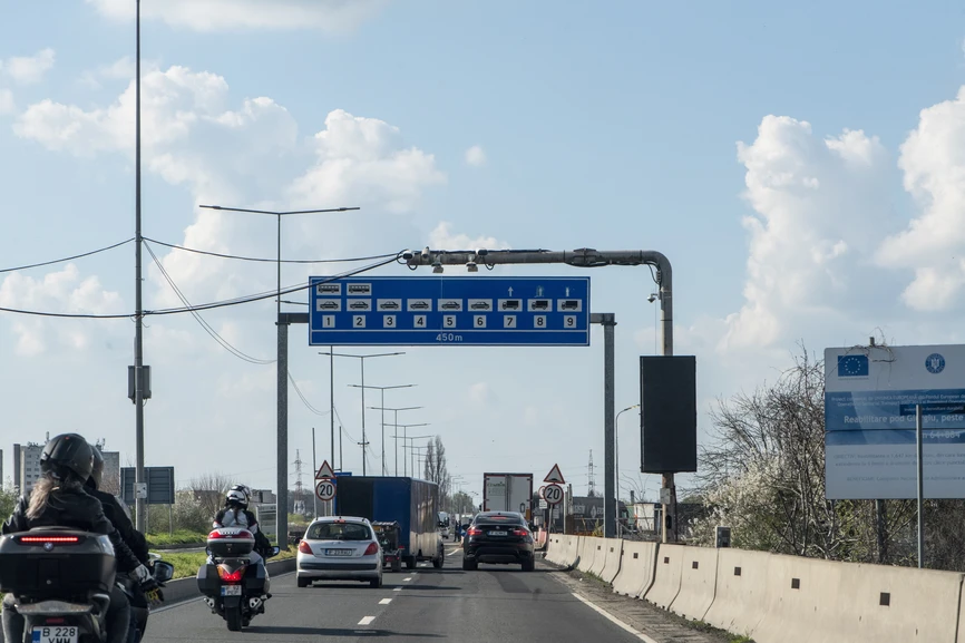 A multi-lane road approach to the Danube Bridge border crossing in Giurgiu, Romania, showing an overhead blue sign indicating lane allocations for buses, cars, and trucks 450 meters ahead, with motorcycles and cars in transit under a bright afternoon sky with scattered clouds, April 2026