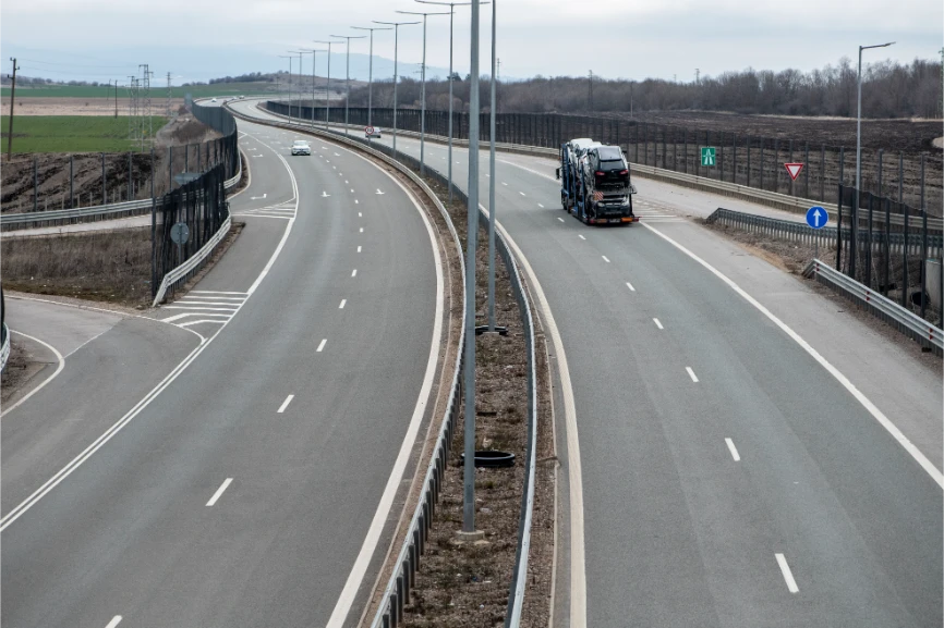 Serbian A1/E75 motorway with light traffic, a car transporter carrying new vehicles on the right carriageway and a passenger car on the left, divided carriageways with anti-glare fencing through open agricultural landscape, March 2026